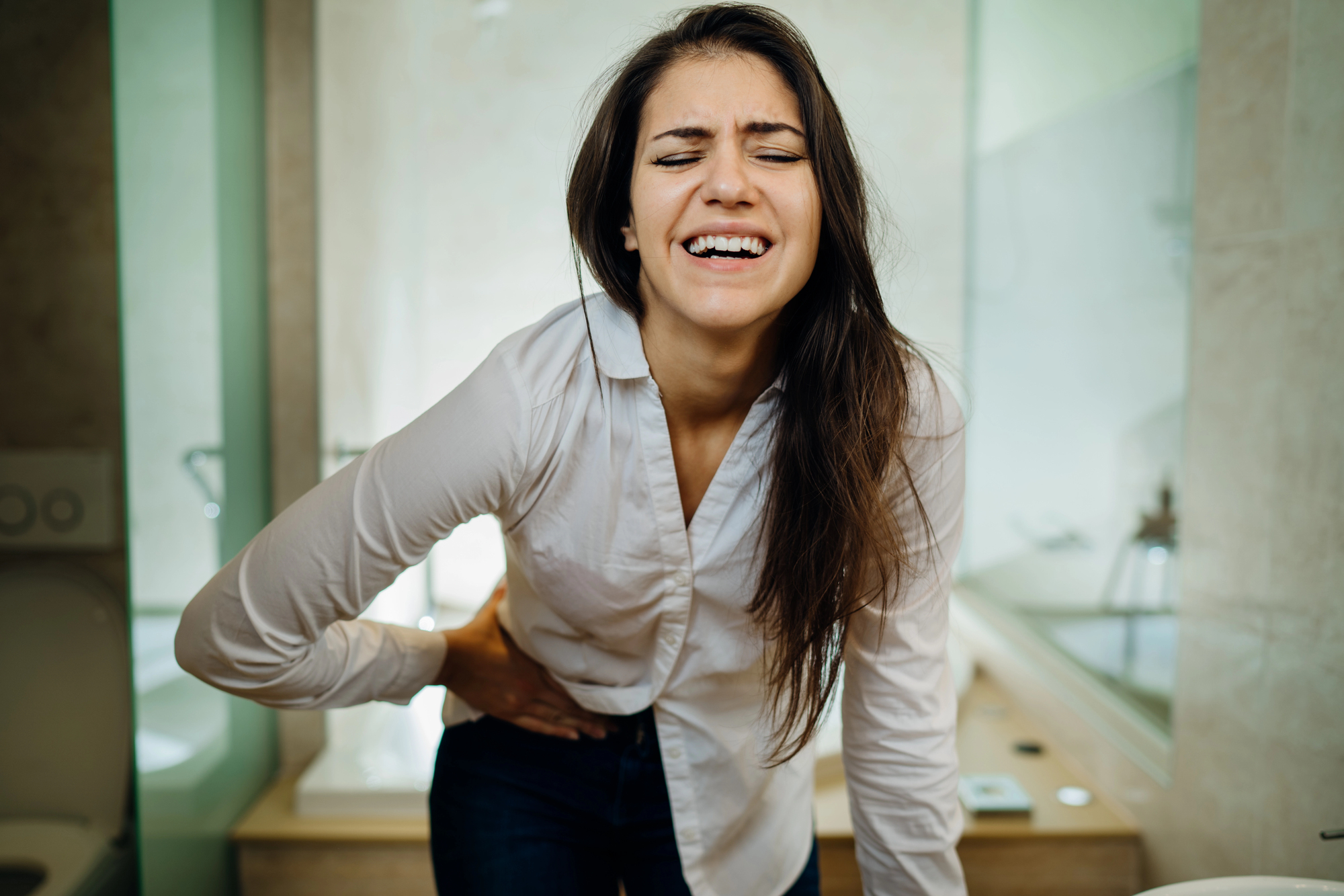A woman clutching her lower abdomen in pain, standing in a bathroom.